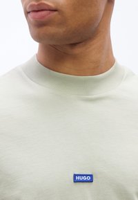 Κοντινό πλάνο ενός ατόμου που φοράει ανοιχτό πράσινο μπλουζάκι με στρογγυλή λαιμόκοψη και μικρό μπλε διακριτικό με λογότυπο "HUGO" στο στήθος.