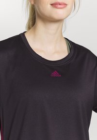 Černé sportovní tričko s kulatým výstřihem a krátkými rukávy, s malým růžovým logem Adidas na hrudi. Hladká, lehká tkanina.