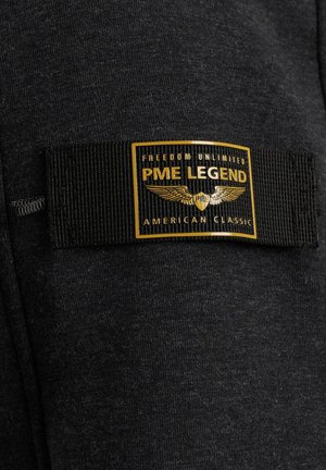 Tissu noir avec une étiquette tissée noire comportant un écusson bordé d'or indiquant "PME Legend", des ailes et "Freedom Unlimited American Classic."