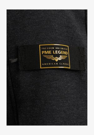 Schwarzer Stoff mit einem schwarzen gewebten Etikett, das einen goldumrandeten Aufnäher mit der Aufschrift "PME Legend", Flügeln und "Freedom Unlimited American Classic" zeigt.