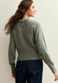 Lichtgrijze trui met een gebreide textuur, ronde halslijn en een sleutelgatdetail aan de achterkant, met versieringen langs de halslijn.