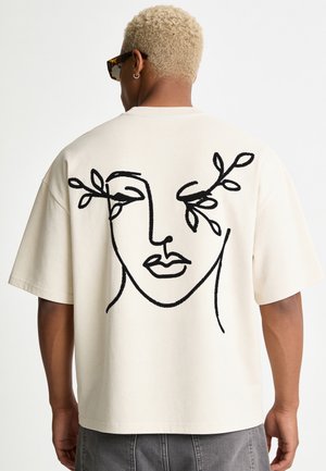 Person mit kurzen blonden Haaren und Sonnenbrille trägt ein weißes T-Shirt mit einem schwarzen abstrakten Gesicht, aus dessen Augen Blätter auf dem Rücken herausragen.
