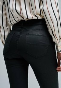 Persona vestida con unos jeans negros ajustados y una blusa de manga larga metida por dentro, con rayas verticales marrones y negras, vista desde atrás.