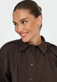 Camicia button-up marrone scuro con colletto, caratterizzata da un design plissettato, texture liscia e dettagli minimi.