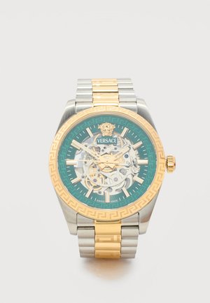 Montre-bracelet Versace avec bracelet en métal or et argent, lunette verte, cadran squelette, index et aiguilles dorés, logo Méduse à 12 heures.