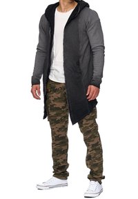 Lange grijze hoodie-jacket met een asymmetrische zoom, gecombineerd met camouflage cargo pants en witte sneakers. Casual stedelijke outfit.