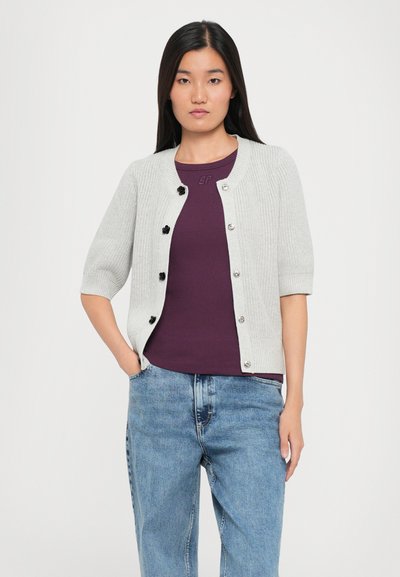 Grå strikket cardigan med korte ermer og svarte knapper, lagdelt over en lilla topp; kombinert med lyseblå, avslappet passform jeans.