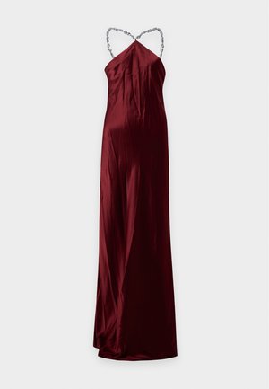 Femme portant une robe en satin rouge foncé sans manches avec un décolleté halter chaîne et des chaussures argentées pointues, debout avec une main sur la hanche.