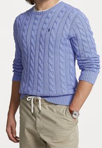 Person som bär en ljuslila kabelstickad tröja med en liten logotyp, beige byxor med dragsko och ett armbandsur med ett brunt läderarmband.