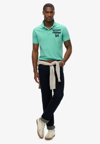 Polo mint avec des lettres foncées, porté avec un jean noir. Pull gris clair noué à la taille et baskets beige.