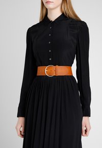 Robe noire à manches longues avec un devant à boutons et une jupe plissée, cintrée à la taille par une large ceinture en cuir beige avec une boucle en argent circulaire.