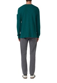 Man in een teal kleurige lange mouwen shirt, grijze jeans en witte sneakers, staande met zijn rug naar de camera op een witte achtergrond.