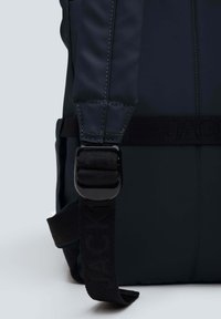 Cinghietto di uno zaino nero con una fibbia in plastica, caratterizzato da dettagli cuciti e una cinghia in tessuto con marchio. Superficie liscia e texturizzata.