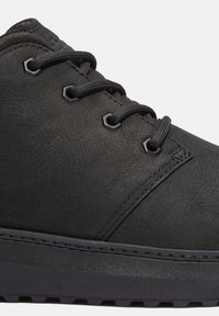 Zapatilla de cuero negro con parte superior texturizada, cordones planos negros, ojetes hexagonales y suela de goma. Diseño minimalista con acentos cosidos.