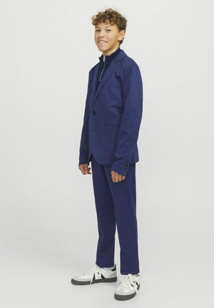 Navy colbert en broek gemaakt van gladde stof. Heeft een enkele knoop, twee voorzakken en een minimalistisch ontwerp, gecombineerd met witte sneakers.