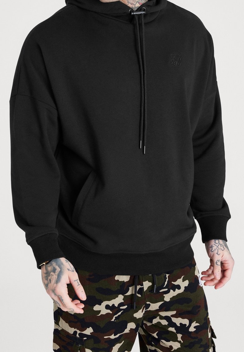 Sweat à capuche noir avec une poche avant, fermeture à cordon et poignets côtelés, assorti à un pantalon cargo camouflage.