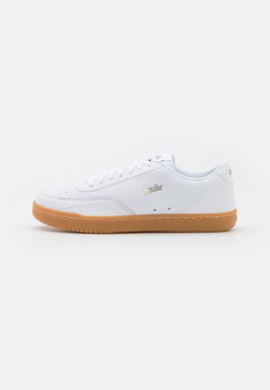 Baskets basses blanches avec semelle en gomme, lacets blancs et petit logo Nike argenté sur le côté, montrées de profil sur fond uni.