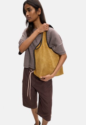 Kobieta ubrana w pasiastą koszulę i brązowe szorty, trzyma na ramieniu dużą beżową zamszową torbę typu tote z czarną lamówką.