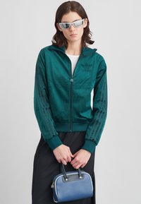 Γυναίκα με τζάκετ Adidas σε τυρκουάζ και μαύρη φούστα, κρατώντας μια μικρή μπλε τσάντα, φορώντας ασημένια φουτουριστικά γυαλιά ηλίου μπροστά από γκρι φόντο.