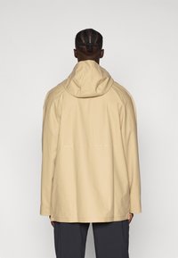 Beige wasserfeste Jacke mit Kapuze, kastigem Design, langen Ärmeln und glatter Textur. Verfügt über minimale Nähte und eine entspannte Passform.