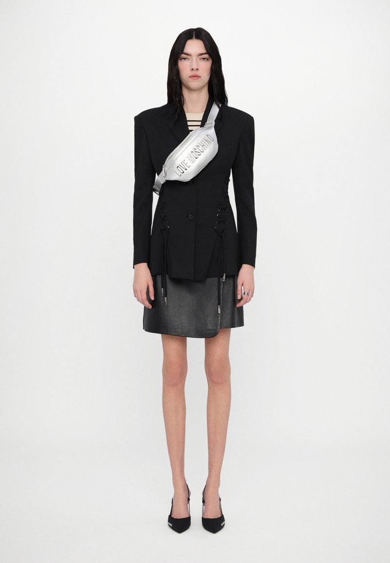 Črni krojeni blazer z strukturiranimi rameni, podrobnostmi za vezanje in oprijetim usnjenim krilom. Srebrna torbica za pas z napisom "LOVE MOSCHINO". Črni petki.