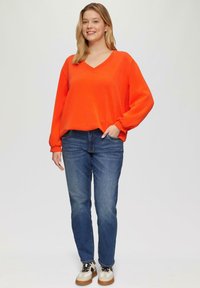Oranges V-Ausschnitt-Pullover mit Ballonärmeln, kombiniert mit dunkelblauen Skinny Jeans und cremefarbenen Sneakern mit schwarzen Schnürsenkeln und braunen Sohlen.