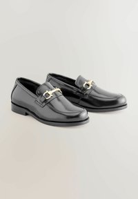 Mocassins en cuir noir avec une finition lisse, arborant un accent en chaîne dorée sur le dessus du pied et une semelle en caoutchouc basse.