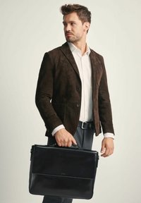 Czarna skórzana teczka trzymana przez mężczyznę w brązowej zamszowej kurtce i białej koszuli. Torba ma klapę z przodu i górny uchwyt.