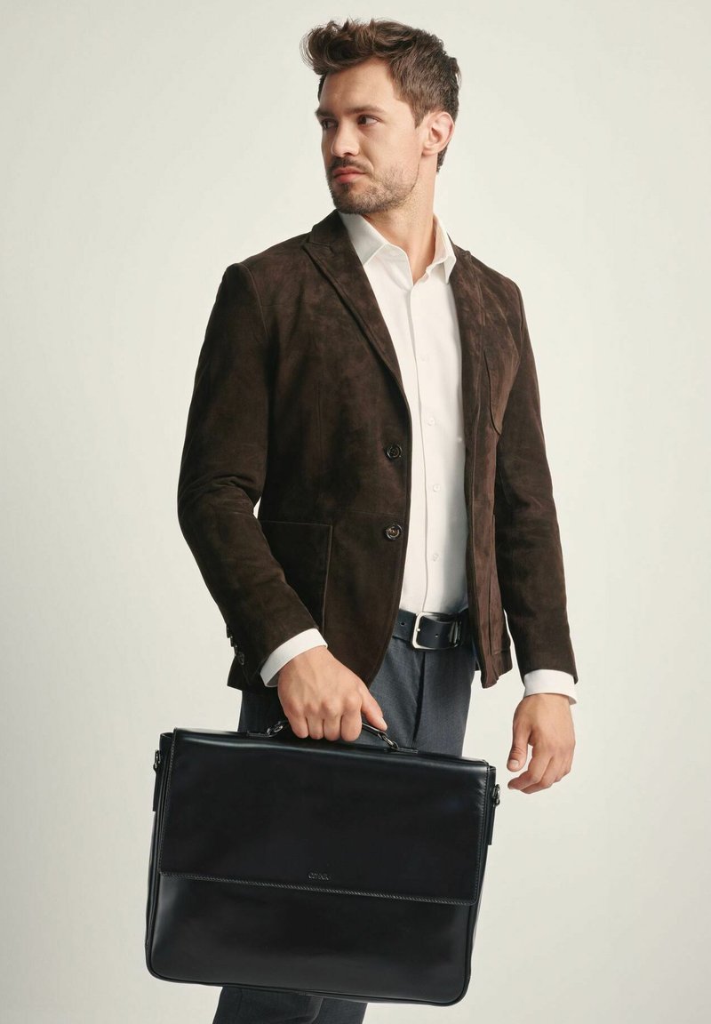 Czarna skórzana teczka trzymana przez mężczyznę w brązowej zamszowej kurtce i białej koszuli. Torba ma klapę z przodu i górny uchwyt.