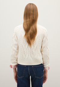 Weißer Strickpullover mit Kabelmustern, geripptem Saum und langen Ärmeln. Kombiniert mit dunkelblauen Jeans und einem rot gestreiften Hemd darunter.