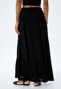Falda maxi negra hecha de tela ligera con volantes escalonados en el dobladillo. Cintura elástica para mayor comodidad. Combinada con sandalias planas de imitación tejido.