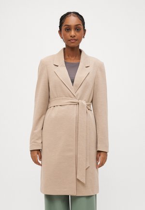Manteau beige ceinturé en tissu texturé, avec des revers nets et de longues manches. Porté sur un haut gris, il présente une silhouette à coupe droite.