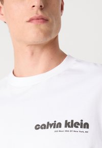 Balts kokvilnas T-krekls ar apaļu kakla izgriezumu un melnu apdrukātu logo, kas lasāms "calvin klein" kopā ar adresi.