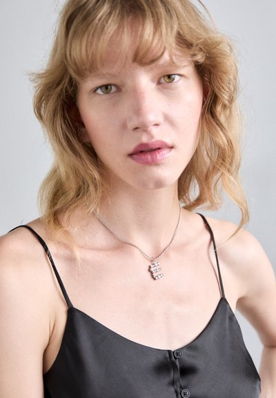 Ασημένιο κολιέ με γεωμετρικό pendant που διαθέτει τρεις σειρές από καθαρές πέτρες. Το μοντέλο φοράει μια μαύρη σατέν τιράντα με λεπτές straps.