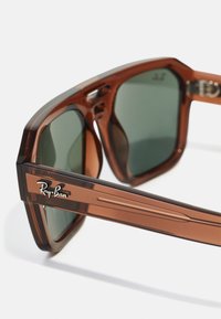 Ochelari de soare din plastic maro cu lentile verzi, cu un cadru gros și accente metalice. Logo-ul 'Ray-Ban' este afișat pe birou.