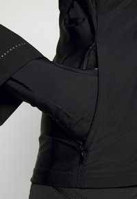 Chaqueta atlética negra con mangas largas, que incluye un bolsillo con cremallera lateral y una textura suave. Puntos reflectantes a lo largo de la manga añaden detalle.