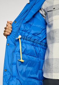 Blauwe pufferjack binnenkant met quilted textuur, elastische tailleband, gele trekkoord en ritszak. Grijs geruit shirt zichtbaar.