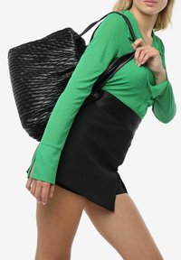 Zwarte geweven handtas met een gestructureerd ontwerp, voorzien van dubbele handvatten, gecombineerd met een groene getailleerde top en zwarte high-waisted shorts.