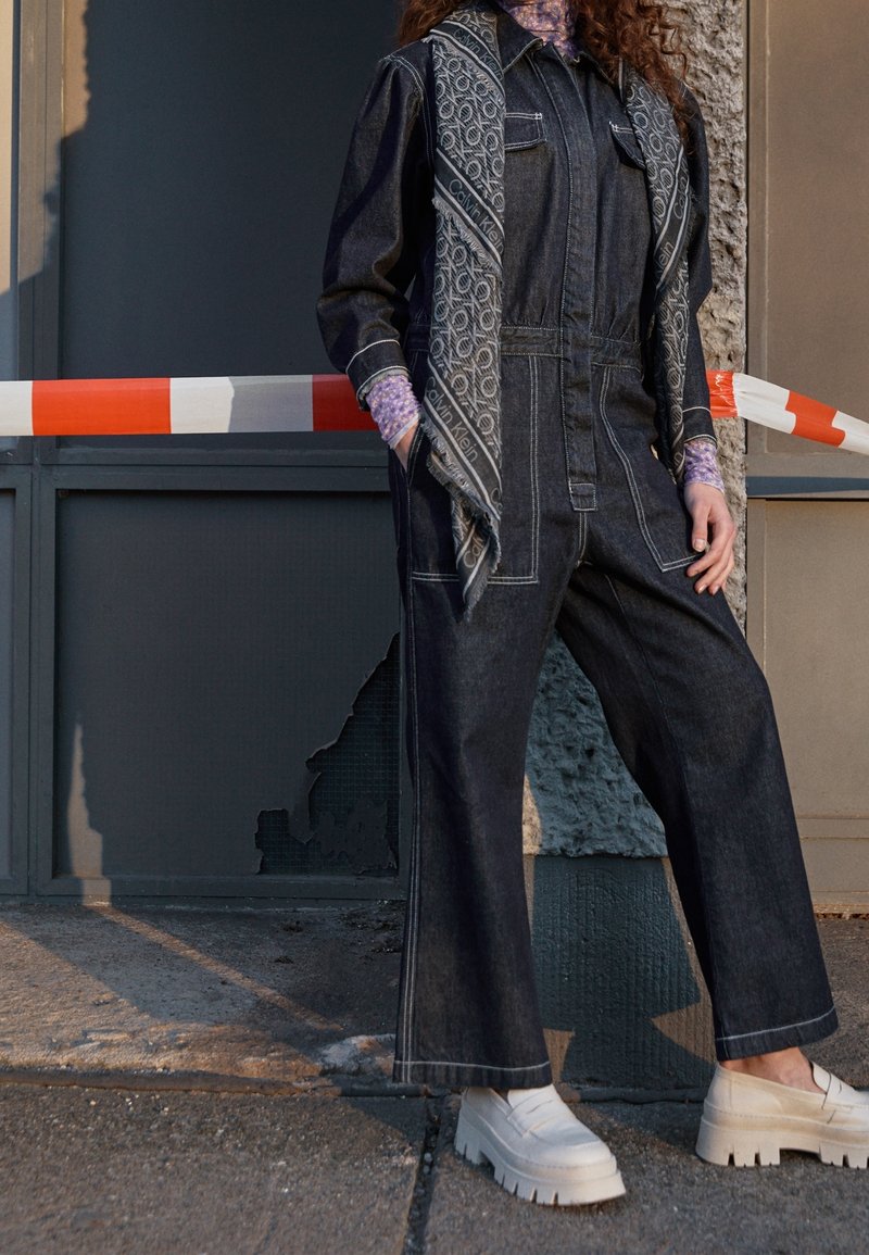 Personne portant une combinaison en denim foncé, une écharpe à motifs et des chaussures blanches épaisses à plateforme, debout près d'un mur gris et d'une barrière orange et blanche.