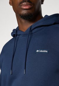 Marineblå hettegenser laget av myk stoff, med snorhette og en liten hvit Columbia-logo på forsiden. Glatt tekstur.