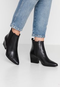 Schwarze Lederstiefeletten mit spitzer Zehe, elastischen Seiteneinsätzen und Blockabsatz. Getragen mit hochgekrempelten hellblauen Jeans.