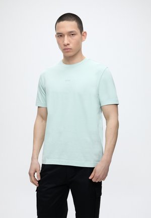 Νέος άνδρας με κοντά μαλλιά, φορώντας ένα ανοιχτό μπλε T-shirt BOSS και μαύρο παντελόνι, στέκεται μπροστά σε ένα απλό λευκό φόντο.