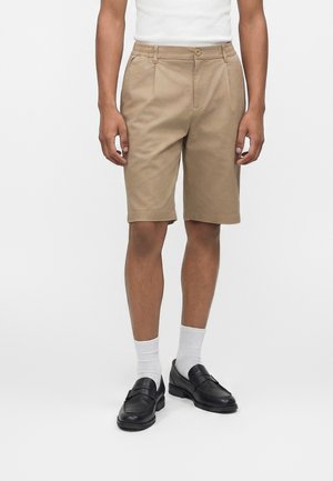 Homme portant un short beige longueur genou, des chaussettes blanches, des mocassins noirs et un haut blanc à manches courtes sur un fond uni.