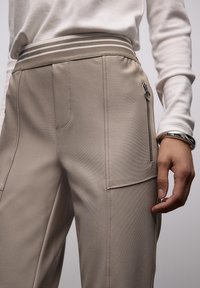 Beige, maßgeschneiderte Hose aus strukturiertem Stoff. Verfügt über eine Reißverschlusstasche und einen kontrastierenden, gestreiften Bund. Hand ruht sanft auf dem Bund.