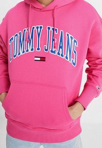 Sudadera de color rosa brillante con "TOMMY JEANS" en letras grandes blancas y azules, y el logo de la bandera de Tommy Hilfiger debajo, con las manos en el bolsillo delantero.