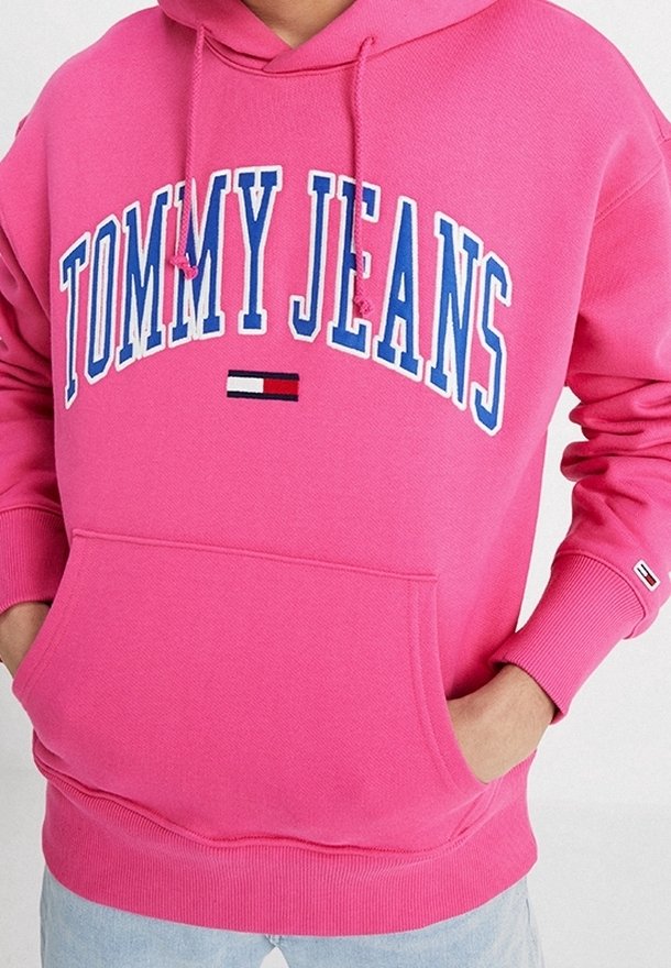 Sudadera de color rosa brillante con "TOMMY JEANS" en letras grandes blancas y azules, y el logo de la bandera de Tommy Hilfiger debajo, con las manos en el bolsillo delantero.