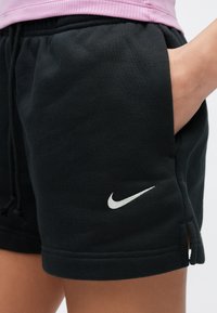Zbliżenie na czarne spodenki Nike z białym logo Swoosh i ręką wsuniętą w prawą kieszeń, noszone z różowym topem.