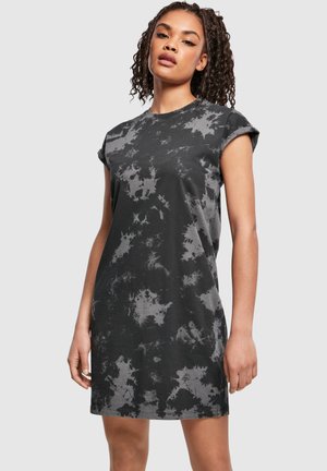 Femeie în picioare, purtând o rochie casual cu mâneci scurte, cu model tie-dye negru și gri, pe un fundal simplu și deschis la culoare.