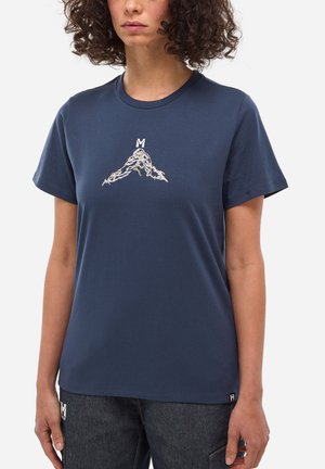 Femme aux cheveux bouclés portant un t-shirt bleu marine avec un graphisme de montagne surmonté d'un "M" blanc, associé à un jean foncé.