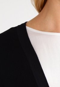 Zwarte gebreide cardigan met een v-halsontwerp, gelaagd over een witte lichte blouse. Glad materiaal, met een eenvoudige, klassieke uitstraling.